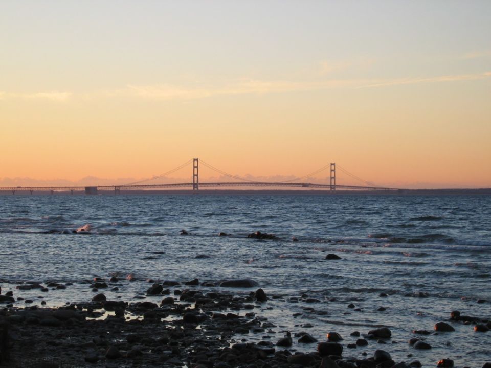Mackinac Bridge spans water under an orange sunset sky, with rocky shore in foreground