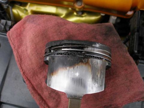 1997 Ford Taurus - A used piston still attached to its connecting rod rests on a red shop rag, showing heavy carbon buildup on the ring lands and a scuffed, s…