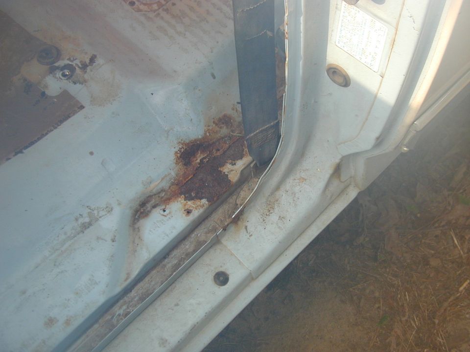 Close-up of rusted interior door frame, light-colored metal, showing corrosion and dirt