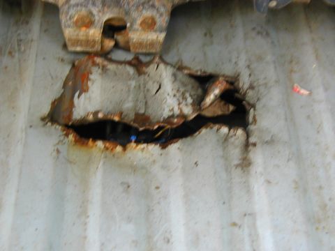 1986 Ford Bronco - A heavily rusted metal panel exhibits a significant hole, revealing damage and internal components. A metal latch is visible at the top.