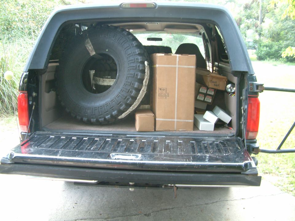 Rear of dark Ford Bronco with tailgate down, cargo area holding a large off-road tire and cardboard boxes