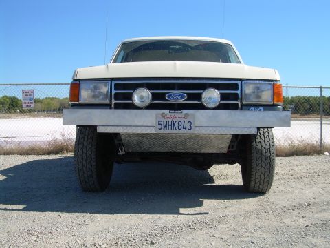 1991 Ford Bronco - Head-on front view of a white Ford 4x4 parked on gravel, wearing a bare diamond-plate aluminum front bumper with two round driving lights m…