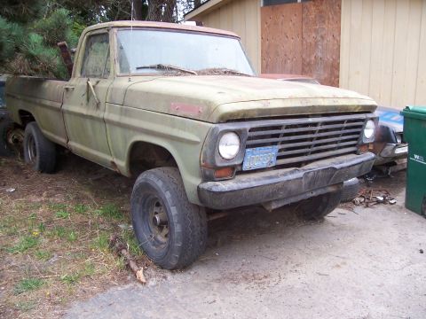 1990 Ford Bronco - This weathered green pickup truck sits in a yard, its front end showing a blue license plate and a prominent grille.