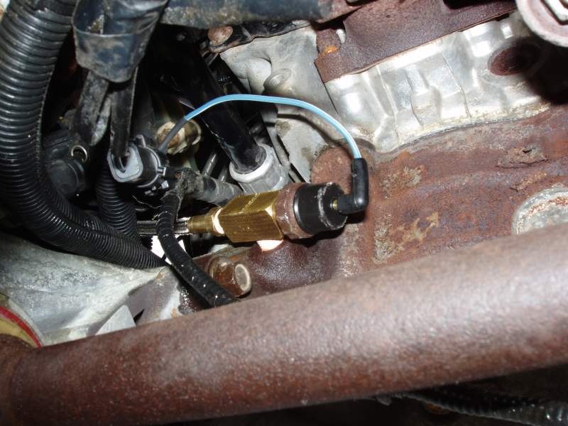 Close-up of a brass sensor with a blue wire connected to engine components and exhaust pipe