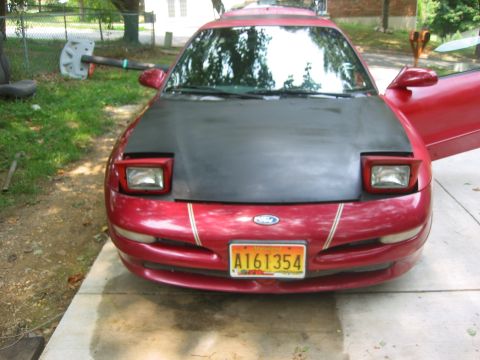 1995 Ford Probe - This red car features a contrasting black hood and distinctive pop-up headlights, with a visible license plate.