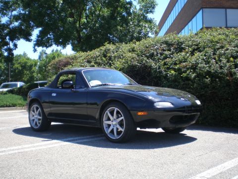 1996 Mazda Miata - A black 1996 Mazda Miata convertible is parked outdoors in front of a building and landscaping.
