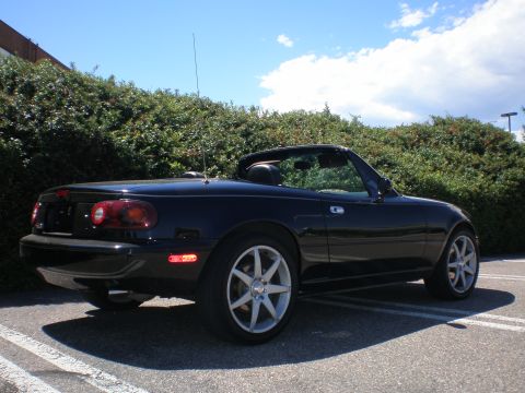1996 Mazda Miata - A black convertible sports car is shown from a rear three-quarter view against a backdrop of green bushes and a blue sky.