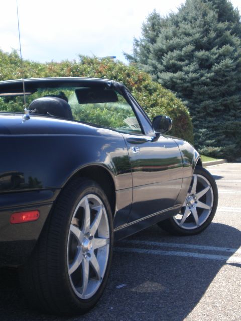 1996 Mazda Miata - A dark convertible sports car is shown from the side, parked in a lot with trees behind it.