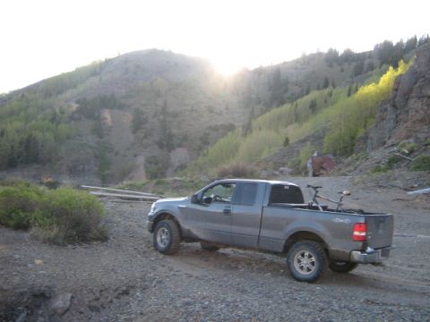 1986 Ford Bronco - Pickup truck with a bicycle in the bed is parked on a gravel road in a mountainous area.
