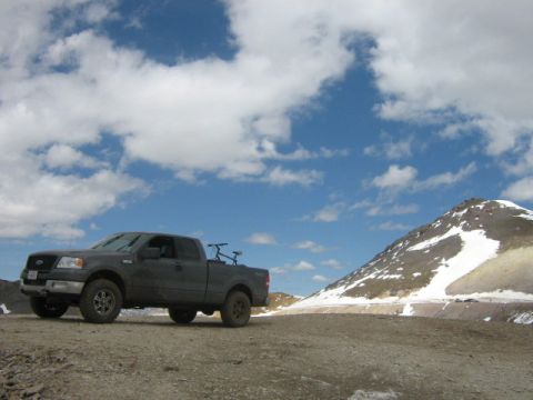 1986 Ford Bronco - A dark gray Ford F-150 pickup truck is parked on a dirt road with a bicycle in its bed, facing a snow-capped mountain under a cloudy sky.