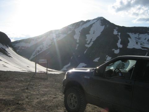1986 Ford Bronco - A dark pickup truck is parked on a dirt road with a snow-capped mountain range in the background under a bright sky.