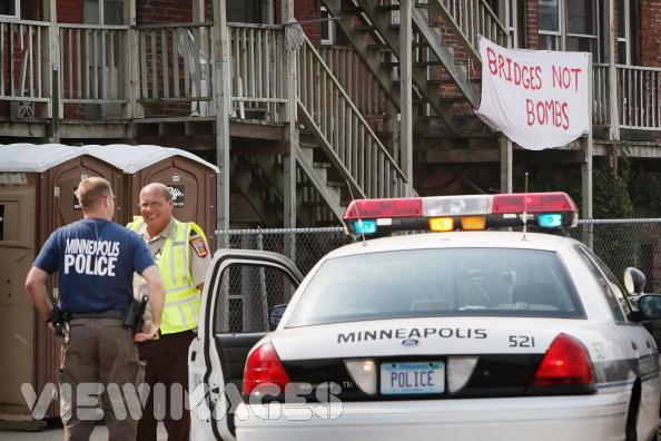 Police officers talking next to a Minneapolis patrol car. Banner reads 'BRIDGES NOT BOMBS'