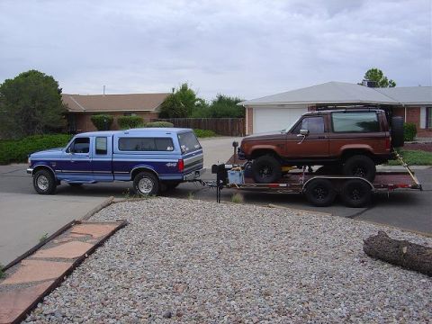 1986 Ford Bronco II - A blue Ford F-150 pickup truck tows a brown Ford Bronco II on a trailer in a suburban driveway.