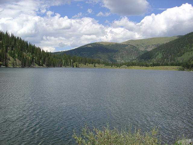 Lake surrounded by pine trees and mountains under a cloudy sky. Water has ripples