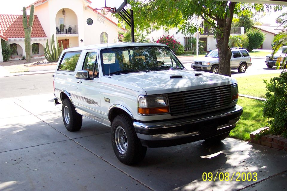White Ford Bronco parked in a driveway. Another SUV and a truck are visible in the background