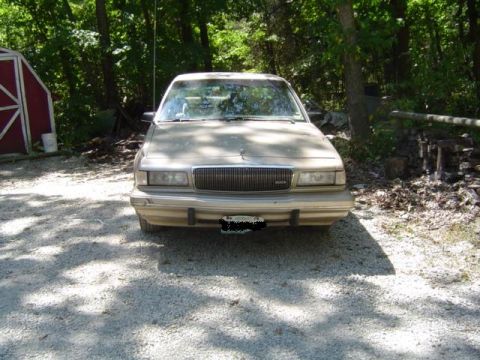 1979 Ford Bronco - Front view of a tan sedan parked outdoors on a gravel surface, with a wooded area and a red shed in the background.