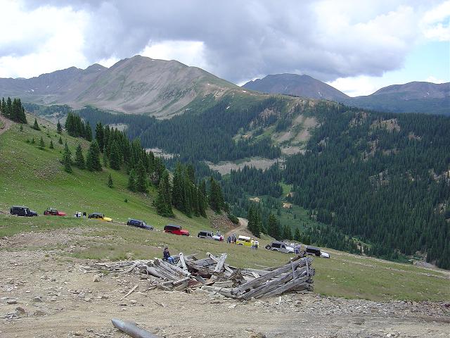 Multiple SUVs driving on a mountain road with pine forests and peaks in the background