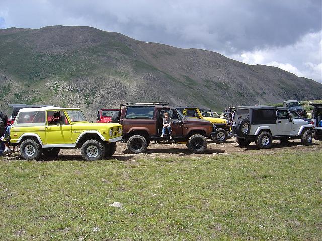 Yellow first-gen Ford Bronco and several Jeeps parked on a grassy hill with mountains in the background