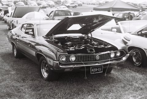 1973 Ford Ranchero - A man in a hat inspects the wheel of a car next to a 1970 Ford Torino GT with its engine exposed.