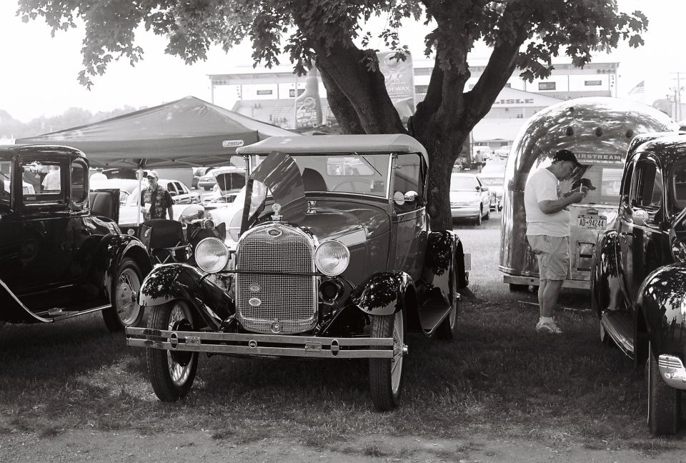 1928 Ford Model A Roadster parked under a tree, with other vintage cars and an Airstream trailer nearby