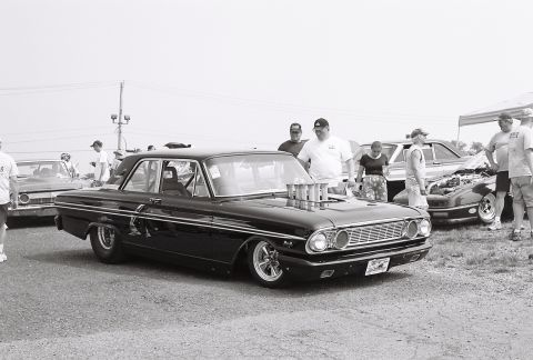 1973 Ford Ranchero - A modified black sedan with a prominent intake system sits on display next to another vehicle with an open hood.