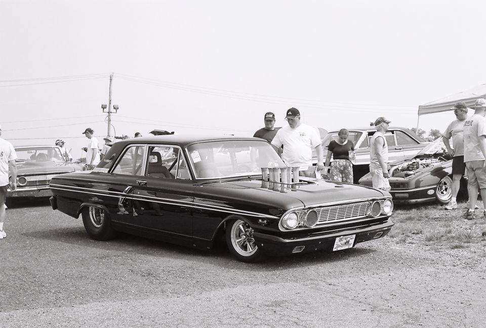 Black sedan with chrome trim and a large intake scoop on the hood. People are gathered around it and another car with its hood open