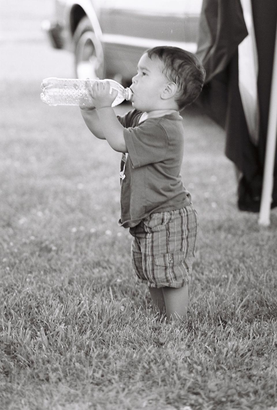 Toddler drinking water from a plastic bottle while standing in grass. Part of a car is visible in the background