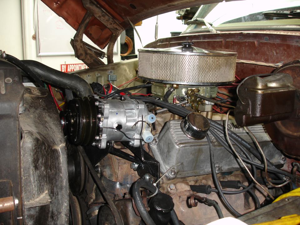 Engine bay showing an aftermarket AC compressor installed on a V8 engine. Air filter housing and carburetor are visible