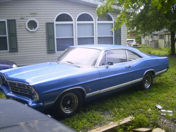 Blue 1967 Ford Galaxie fastback parked on grass in front of a tan house, front three-quarter view