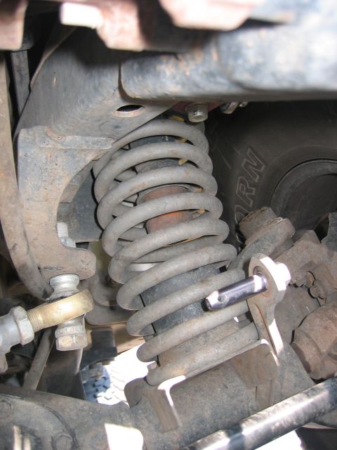2001 Jeep Wrangler - Tight underbody close-up of a dusty coil spring seated on an axle spring perch, with the shock body inside the coil, a tie-rod end and adju…