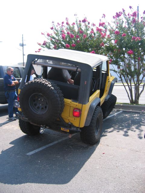 2001 Jeep Wrangler - Rear three-quarter view of a yellow Jeep Wrangler TJ with doors removed and the soft top up, parked in a sunny lot beside blooming crepe my…