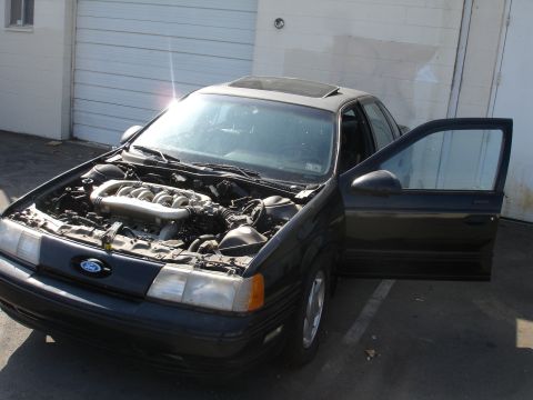 1991 Ford Taurus - A dark sedan with its hood open and driver's door ajar, revealing the engine and a sunroof.