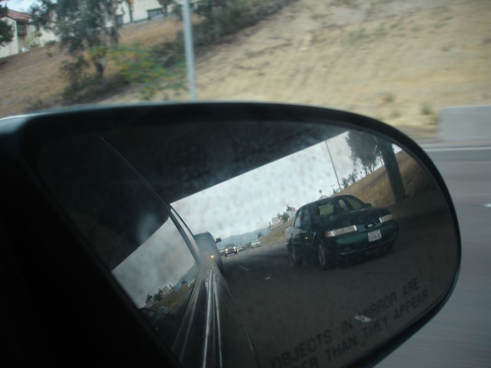 Dark green sedan, possibly a 1992 Ford Taurus, seen in side mirror. License plate visible