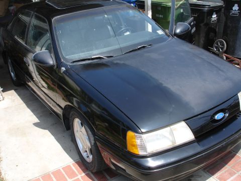 1991 Ford Taurus - A black sedan with a sunroof is parked outdoors, showing its front grille with a Ford emblem.