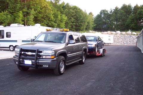 2001 Ford Crown Victoria - A grey Chevrolet Suburban tows a dark blue sedan on a trailer, with RVs and storage units in the background.