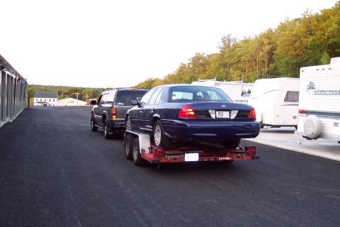 2001 Ford Crown Victoria - A blue 2001 Ford Crown Victoria is transported on a trailer, followed by a dark SUV and other vehicles.