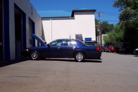 2001 Ford Crown Victoria - A blue 2001 Ford Crown Victoria is parked outside a building with its hood up, with other vehicles visible in the background.