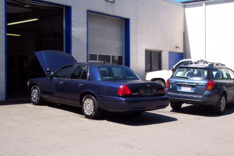 2001 Ford Crown Victoria - A dark blue 2001 Ford Crown Victoria is parked outside a garage with its hood up, alongside other vehicles.
