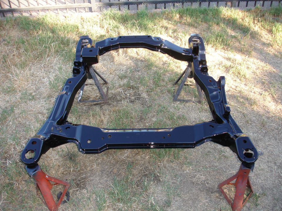 Glossy black restored subframe crossmember on jack stands in a grassy yard.