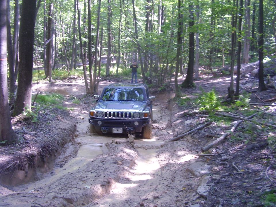 Hummer H3 driving through mud on a wooded trail. A person stands on the roof