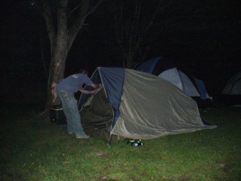 1991 Ford Probe - A person is busy setting up a tent in a grassy area at night, with several other tents visible.