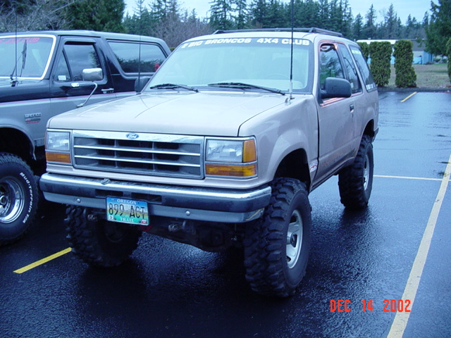 Tan lifted Ford Explorer two-door on mud tires with 4x4 club windshield banner, wet lot