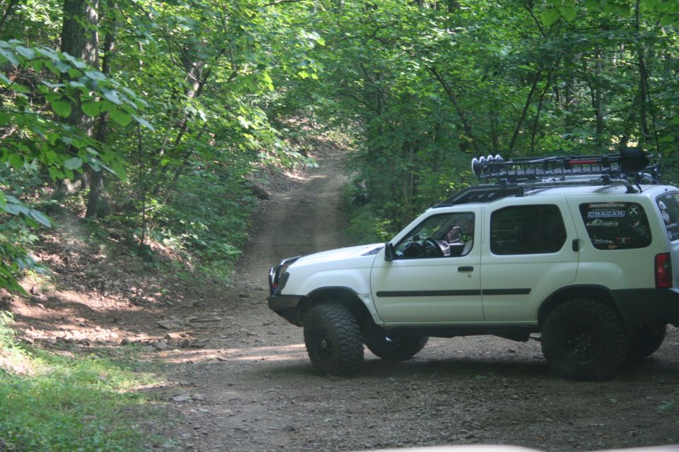 White Nissan Xterra with roof rack and off-road tires on a dirt trail in the woods