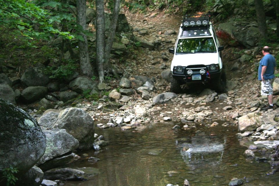 White Nissan SUV with roof lights and bull bar crawling down a rocky trail toward a creek, spotter standing nearby