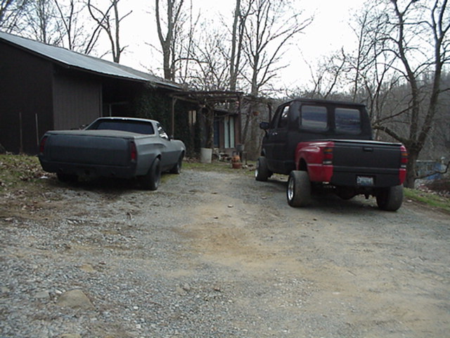 Gray Ranchero and red pickup parked rear-view on gravel drive