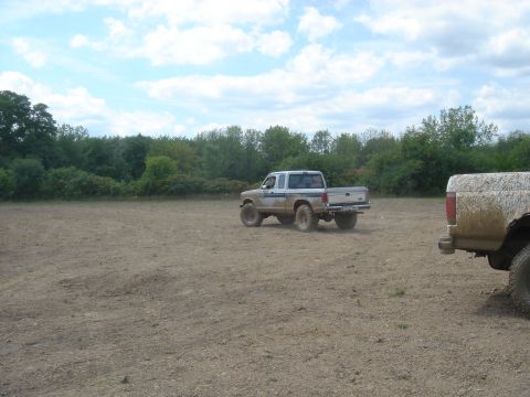 1979 Ford F-250 - Two trucks are seen driving on a dirt field with trees in the background under a cloudy sky.
