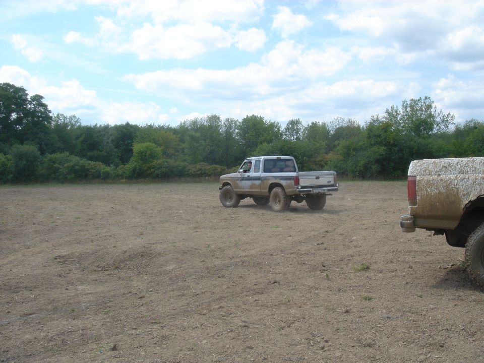 Mud-covered white and blue Ford truck driving on dirt, rear view. Another mud-covered truck is partially visible on the right