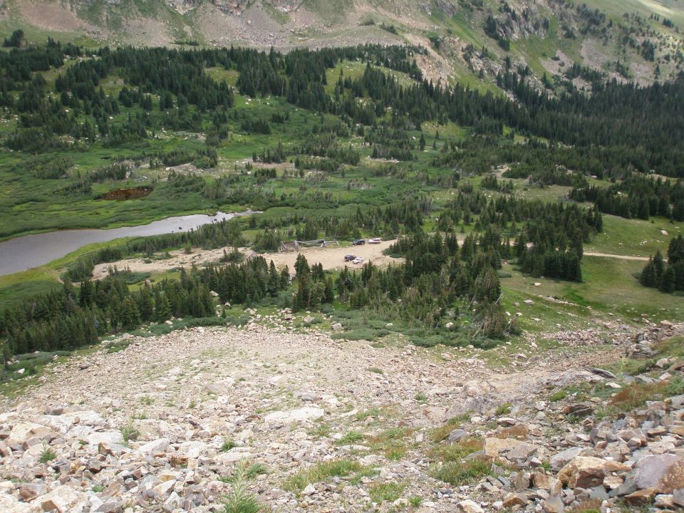 Mountain valley with forest, pond, and tiny distant vehicles on a dirt clearing