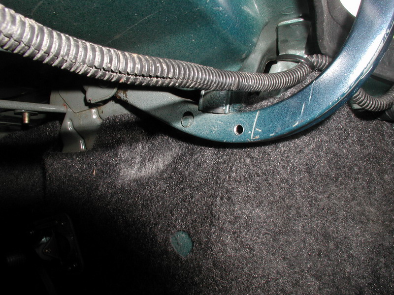 Freshly drilled holes and layout marks on teal trunk body seam beside wiring loom and hinge
