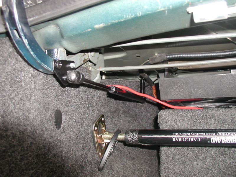 Trunk hinge and gas strut connection above gray carpet with a Highland cargo bar